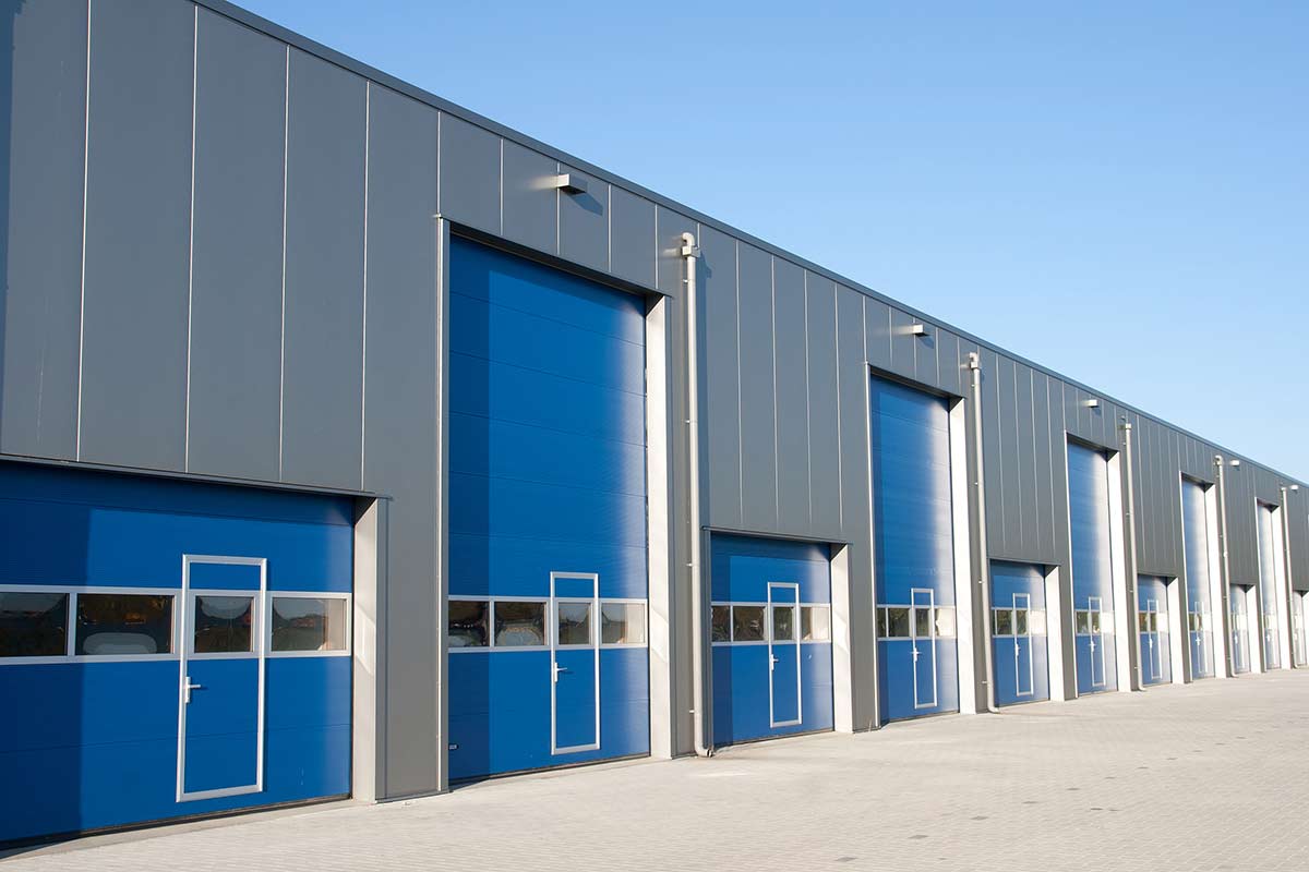 a storefront with mutiple types of commercial doors. its a commercial front with regualr size garage door next to a tall coiling door. The building exterior is gray and the doors themselves are blue.